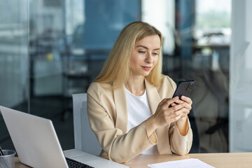 Successful beautiful and happy businesswoman working inside office at workplace, blonde holding phone using online application, smiling sitting at table with laptop, satisfied with work achievement