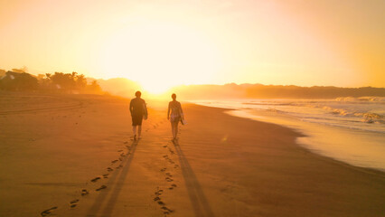 Fototapeta na wymiar SILHOUETTE: Surf couple walking along gorgeous sandy beach towards golden sunset