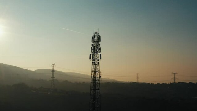 4K drone shot of telephone mast of cell tower. Aerial view of telecom antenna and cellular base station against sunrise and small town surrounded by forests and hills during golden hour stock video.