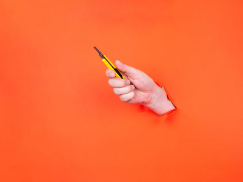 A man's hand holds a yellow cutter through torn orange paper. Hand with sharp knife.

