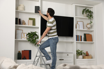 Man on metal folding ladder taking book from shelf at home