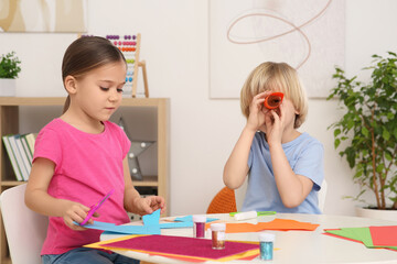 Cute children making paper toys at desk in room. Home workplace