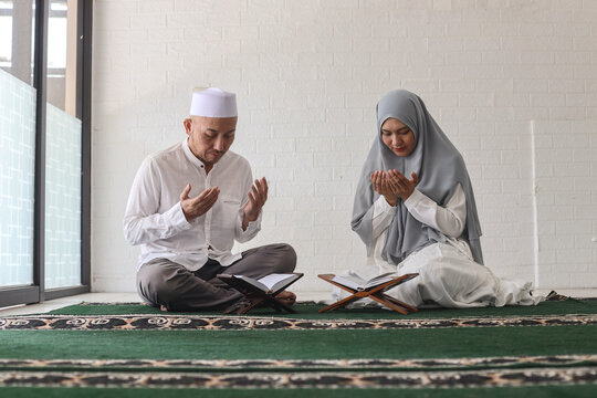 Muslim man with skullcap and woman wearing traditional hijab siting on the prayer mat and raising arms make a dua in mosque together