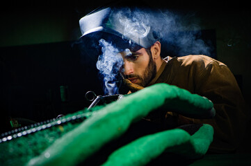 A man in a welding helmet inspects a smoking weld on a piece of metal and concrete sculpture