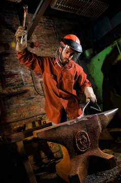 A man wearing a maroon work shirt and a safety mask hammers a piece of rebar on an anvil in a metal shop