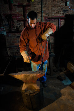 A man wearing a maroon work shirt and leather gloves hammers a piece of rebar on an anvil in a metal shop