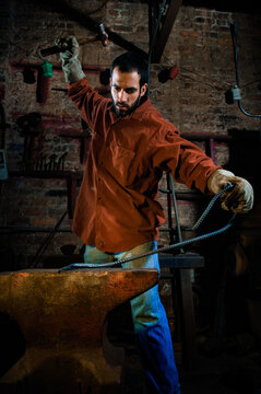 A man wearing a maroon work shirt and leather gloves hammers a piece of rebar on an anvil in a metal shop