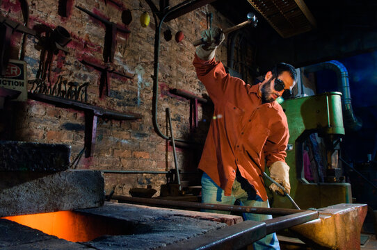 A man wearing a maroon work shirt, leather gloves and safety goggles hammers a piece of rebar at a forge