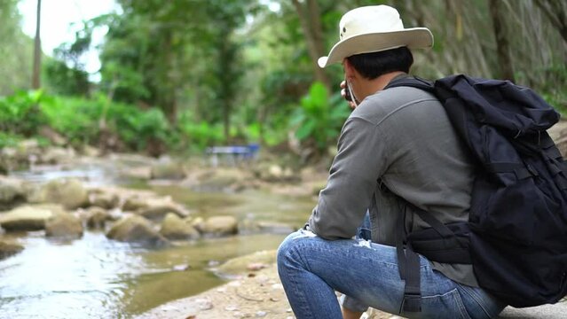 A young man who enjoys trekking for a leisure holiday rests. Pause at the edge of a small stream and bring cell phone to take pictures of the surrounding natural beauty of the forest.