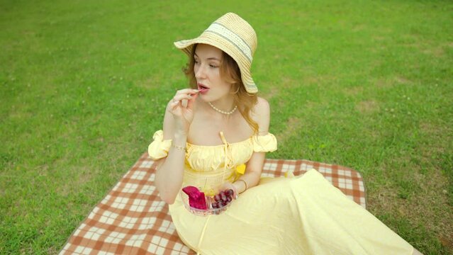 Young woman in yellow sundress enjoying picnic in park eating fruit and reading.