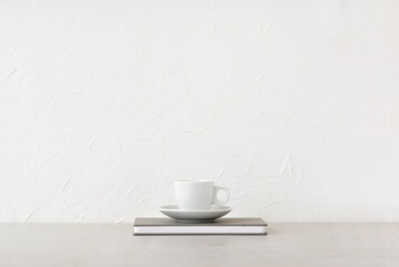 Fototapeta na wymiar White cup with drink and notebook on neutral beige table, white concrete wall background with empty copy space. Minimalist brand template, coffee break, business lunch concept, home office workspace