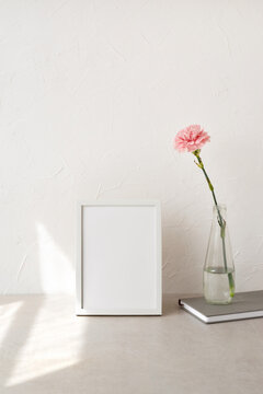 Empty white picture frame, art poster mockup, vase with carnation flower, notebook on neutral beige table and concrete wall background, aesthetic minimalist home workspace with sunlight pattern