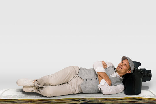 Smiling lazy male photographer relaxing on dollars money stack isolated on white background, Grinning male lensman enjoying a leisurely moment while lounging on a stack of currency.