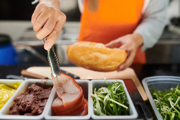 Focus on metallic tongs held by young female clerk taking slice of fresh bacon before putting it into hotdog while making fast food