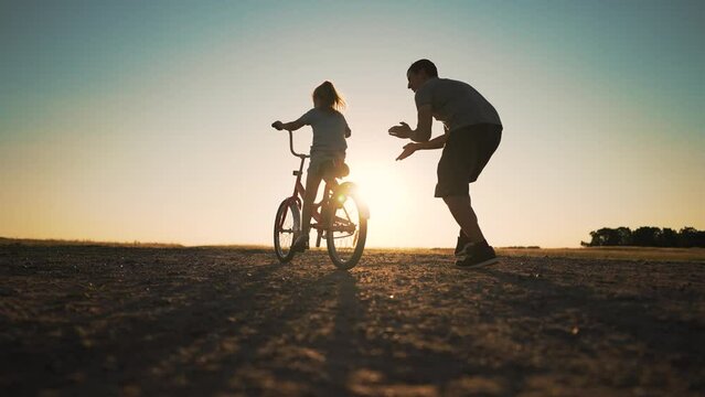 Happy family green park at sunset.Dad teaches daughter to rid bike in summer park.kid rides bicycle on green grass.Green energy.Dad teaches his daughter to ride bike for first time in park at sunset.