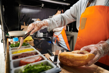 Gloved hands of young woman with metallic tongs taking slice of bacon from container before putting it into fresh bun while preparing hotdog - obrazy, fototapety, plakaty
