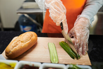Hands of woman in protective gloves slicing fresh cucumber on wooden chopping board while preparing vegetarian hotdog in food truck
