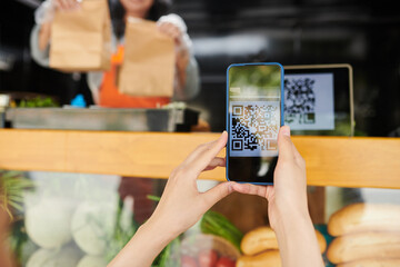Hands of young female customer scanning qr code of products on smartphone while buying takeaway street fast food in truck