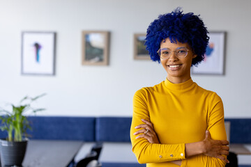 Portrait of biracial casual businesswoman with blue afro and glasses smiling in office, copy space