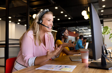 Happy caucasian casual businesswoman and diverse colleagues using computers and headsets in office