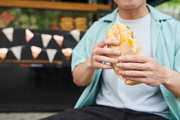 Close-up of young hungry man in casualwear holding appetizing hotdog in paper while standing in...