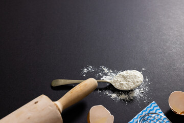 High angle view of flour in spoon with rolling pin and eggshells on black background, copy space