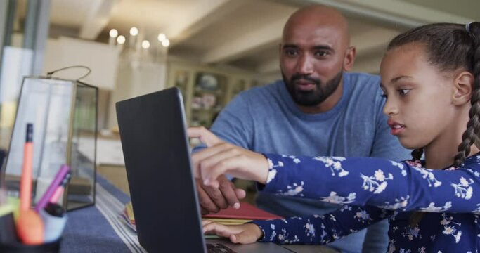Happy biracial father and daughter learning online using laptop at table, slow motion