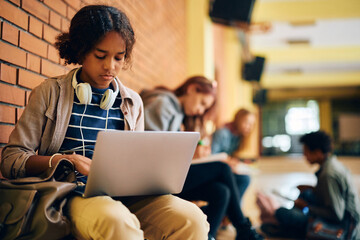 Black teenage girl e-learning on laptop in hallway at high school.