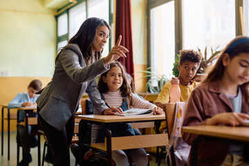 Hispanic female teacher assists schoolgirl with new lesson in classroom.