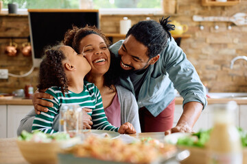 Loving black family enjoys while having meal together at home. Daughter is kissing her mother.