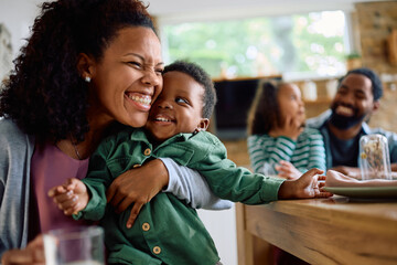 Obraz premium Cheerful black mother hugs her son tightly during family meal at dining table.