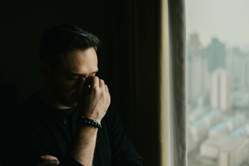 Frustrated man stands by a window and puts his hand to his face