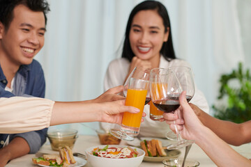 Hands of young intercultural friends clinking with wineglasses with red wine and glasses of orange juice over served table against happy couple