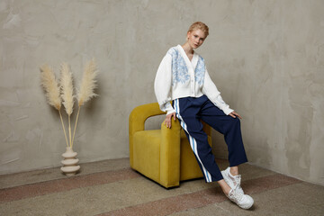 Fashion photo of beautiful elegant young woman in a pretty white sweater, sport pants, trousers with the stripes posing over sandy beige textured wall. Studio Shot. Blonde model sits on a yellow chair