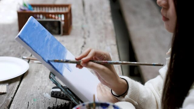Close-up woman's hand using long-handled paintbrush, paint water in bright blue color, paint color sky on top white sheet, is blending colors even out all colors make picture even more beautiful.