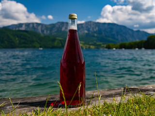 a bottle with a red drink stands on a wooden board against the backdrop of a lake and mountains