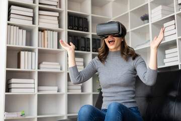 Surprised young latin woman in VR headset touching the air. Smiling caucasian woman wearing vr glasses virtual reality technology device watching 3d simulation video while sitting on sofa.