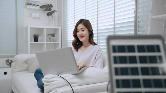 Asian woman using computer laptop surfing website and internet on sofa in living room have solar panel on table give energy in home. Alternative energy and clean energy concept