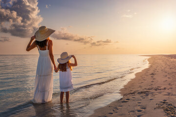 A mother and her daughter in white summer clothing standing on a beautiful beach and enjoying the summer sunset