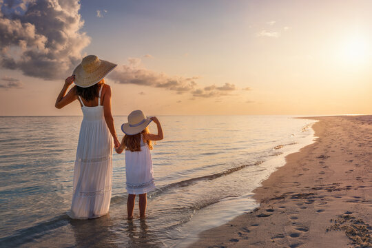 A mother and her daughter in white summer clothing standing on a beautiful beach and enjoying the summer sunset