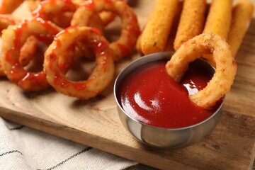 Tasty fried onion rings, cheese sticks and ketchup on wooden board, closeup