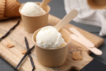 Paper cups with delicious ice cream and vanilla pods on wooden board, closeup