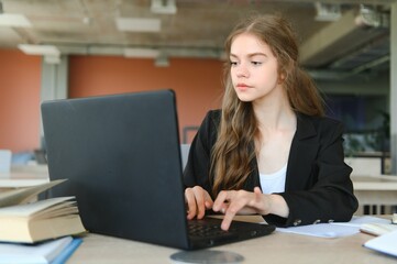 A beautiful caucasian female student is studying in college remotely. She is sitting with a laptop and a notepad and concentrated is watching a video conference lesson