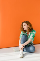 Full length of young red haired woman in modern blouse with floral pattern and jeans sitting on...