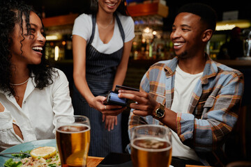 Happy multiracial friends in casual clothing paying waitress for dinner and drinks at restaurant