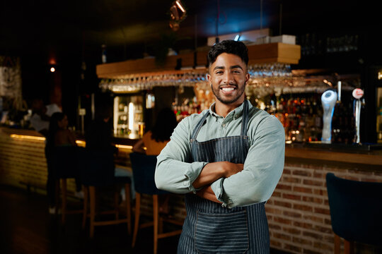 Young biracial man in apron smiling with arms crossed while working as waiter in restaurant - Powered by Adobe