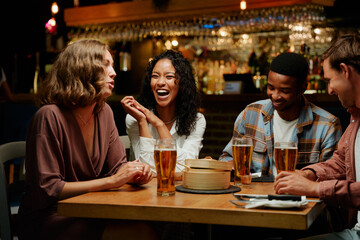 Happy young multiracial group of friends in casual clothing talking and laughing over dinner at bar
