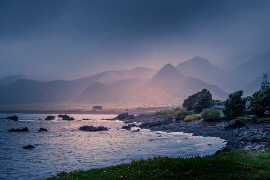 Rain clouds low over rugged green hills surrounding black sand beach. Stormy day at Cape Palliser, North Island, New Zealand