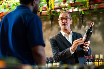 Professional African man wine shop worker explaining and recommending wine to customer at liquor...