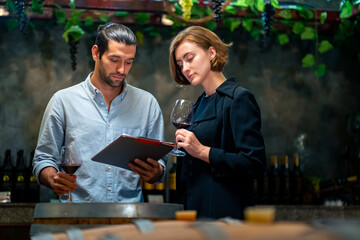 Professional man and woman sommelier testing red wine in wine glass with tasting and smelling at wine cellar with wooden barrel in wine factory. Winery manufacturing industry and winemaker concept.